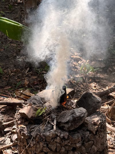 The image depicts a smoking fire in an outdoor environment, possibly in Moo 7, Thailand. A metal grate surrounds a pile of rocks and debris, within which a small fire is burning. Thick white smoke emanates from the fire, obscuring some of the surrounding area. The ground is covered in leaves and other natural materials. A green plant is visible in the foreground.