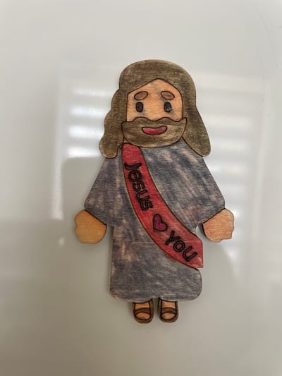 The image shows a wooden cutout of Jesus Christ, depicted as a cartoonish figure with brown hair and beard, a grey robe, and brown sandals. A red sash with the words "Jesus loves you" and a heart symbol is draped across his chest. The cutout is placed on a white, reflective surface, likely a table or countertop. There are no visible contextual clues to indicate its location in the city of Cahul, Moldova.