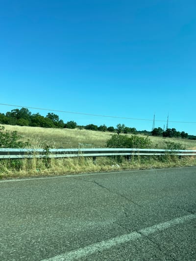 The image displays a roadside scene in Cassano delle Murge, Italy, under a clear, bright blue sky. The upper third of the frame is dominated by the unobstructed blue sky. Below the horizon, a distant line of dark green trees is visible, with a gentle slope of dry, golden-brown grass and scattered green shrubs extending into the midground. Two tall, lattice-structured communication or utility towers stand on the right side of the frame, rising above the tree line. A thin, dark utility wire traverses horizontally across the upper midground.

In the lower midground, a two-tiered silver metal guardrail extends diagonally from left-center to right-center, separating the grassy area from the paved road. The ground immediately adjacent to and behind the guardrail is overgrown with dry grass and weeds, and a piece of white debris is visible near the guardrail on the right side. The lower section of the image reveals a dark asphalt road surface, exhibiting visible cracks and featuring a faded white solid line marking its right edge. The road and guardrail show slight motion blur, consistent with an image captured from a moving vehicle. The scene is devoid of any individuals, vehicles, or animals.