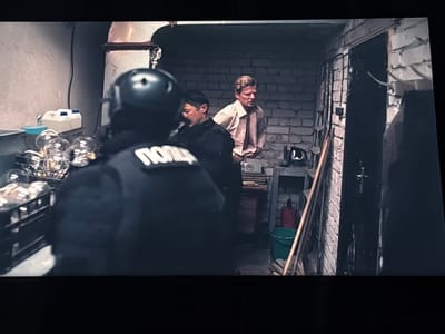 The media file depicts an indoor scene, likely a dimly lit room or basement, with rough brick walls. Three individuals are visible. In the foreground, facing away from the camera, is a person wearing a dark tactical vest over a black uniform and a black helmet. White Cyrillic text, reading "ПОЛИЦ" (partially visible), is printed on the back of the vest. To the right and slightly behind the first person, another individual, appearing to be male, is seen from the back, wearing a dark jacket. Further to the right and in the background, a fair-haired man in a light-colored collared shirt is standing, looking towards the other two.
The room contains several objects. To the left, a shelf holds containers, including what appear to be two large plastic jugs and a collection of clear, lightbulb-like objects. Further to the right, a wooden shelf unit against the brick wall holds various items, including a red canister and a green bucket. A metal pipe runs vertically along the wall behind the first individual. The overall atmosphere suggests a tense or clandestine situation, possibly an interrogation or arrest, given the presence of the person in tactical gear. The lighting is dim, contributing to a somewhat gritty and enclosed feel. There are no clear indications of time of day or weather.