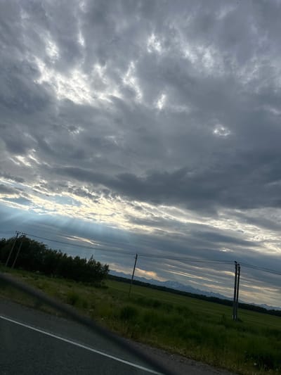The image captures a landscape scene under a cloudy sky with visible sunbeams. In the foreground, a section of a road with a white line is visible, angled from the bottom left corner. To the right of the road, a green grassy field stretches into the distance. A line of trees borders the field on the left. In the background, mountains with snow-capped peaks are discernible. Two utility poles with wires are present in the middle ground. The sky is filled with dramatic, layered clouds, with patches of sunlight breaking through, creating shafts of light that illuminate the scene. The location is identified as Кеткино, Russia.