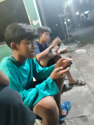 In Barengkrajan, Indonesia, at night, two young boys are focused on their mobile phones, seated on a concrete step. The boy in the foreground, wearing a teal long-sleeved shirt, is captured from the chest up, his head turned to his right. Beside him, slightly behind and to the right, a second boy in a blue shirt is also engrossed in his phone, his face partially obscured. The ground is paved with concrete, and the background is dimly lit, suggesting an outdoor, possibly residential area, with some foliage and structures visible. The overall atmosphere is casual and focused on individual digital engagement.