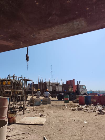 This image depicts an outdoor industrial scene in the city of Chimbote, Peru, likely within a shipyard or similar facility. The foreground is dominated by a large, rusty, red-brown metal structure, possibly the underside of a ship or dock. Below this, a thick, frayed rope hangs from the structure.
In the middle ground, a group of individuals are engaged in work. Two men in hard hats, one yellow and one orange, stand near a large industrial truck. The truck has a red cabin and a trailer filled with wooden poles and other materials. Several individuals can be seen working on or around scaffolding structures. To the left, a large stack of nets with yellow buoys is secured to a metal frame. Various metal barrels, including one green with a "Castrol" label, are scattered around the area, some upright and others overturned. Piles of debris and refuse are visible on the ground, suggesting a working or disused industrial area.
The background is characterized by a clear blue sky. In the distance, a tall antenna tower with multiple levels is visible. The ground surface appears to be a mixture of dirt and gravel. The overall impression is one of a busy, working environment with signs of industrial activity and material handling.