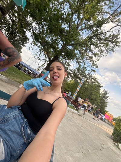 The image depicts a low-angle, wide-angle outdoor selfie featuring a young adult female in the foreground, situated in Kostievo, Bulgaria.
The primary subject, a light-skinned female with dark hair pulled back, occupies the lower-left to mid-right of the frame. She wears a black spaghetti strap top and blue denim shorts. A silver-colored necklace with small charms is visible on her neck. Her left hand, covered by a light blue disposable glove, is raised, forming a "V" or peace sign with her index and middle fingers. Her tongue is extended from her mouth. Part of her right upper arm, adorned with a detailed tattoo, is visible in the upper-left corner, suggesting another individual is seated closely beside her. She appears to be seated on a textured mat or patterned surface.
The mid-ground to background reveals an outdoor event or gathering. To the left, a blue tent structure is visible, surrounded by several individuals sitting and standing. A Bulgarian flag (white, green, red stripes) is partially visible near this tent. Large, mature trees with dense green foliage dominate the upper portion of the image, providing shade. The sky above is partly cloudy with patches of blue.
To the right, a paved pathway extends into the distance. A multi-directional signpost with numerous colored signs is positioned near the trees. Several people are scattered along the pathway, including a man in a yellow and black striped shirt near a market stall, and further down, a man walking with a small child. In the far right background, a bright pink inflatable structure, possibly a bouncy castle, is discernible. The overall scene suggests a local fair, market, or community event taking place on a bright day.