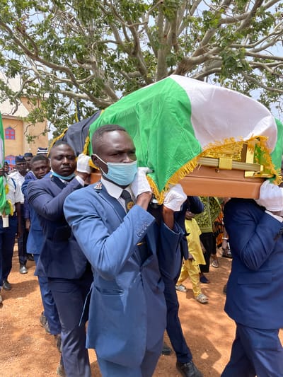 A group of men wearing light blue military uniforms are carrying a wooden coffin draped with the Nigerian flag. The men in the foreground wear light blue surgical masks. The ground is red. Some are wearing black shoes. The location is a red dirt area in Ladura Foron, Nigeria. A tree is on the left. More people stand behind the coffin carriers. Some onlookers wear yellow clothing.