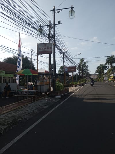 The image is a street scene in what appears to be a developing area. The road is asphalt and has a single solid white line marking the shoulder. A person rides a scooter down the road. Power lines dominate the upper part of the frame, creating a tangled web across the sky.

A roadside restaurant called "Taman Sari" is visible on the left, with signage in English and possibly Indonesian. A flag is displayed in front of the restaurant. There are people sitting at tables near a small food stall, sheltered by a green and red umbrella. There is an advertisement that says "Restauran Taman Sari". The location in the city of Nhot Chang, Laos is unconfirmed.
