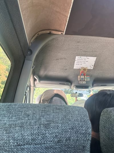 This image captures the interior of a vehicle, likely a marshrutka or bus, in Kholodnaia-Rechka, Abkhazia. The camera is positioned from a passenger's seat looking forward and upward. The ceiling of the vehicle is covered in a textured, light brown material, with a section of darker material and a strip of LED lights visible on the far right.
Affixed to the ceiling is a white sign with handwritten text in Cyrillic script, listing various items and prices, possibly for sale or services. Below the sign, a cluster of religious icons is suspended. These icons appear to be small, framed images of religious figures, possibly a common practice in some regions. A few small twigs or dried plant material are also intertwined with the icons.
In the foreground, the backs of passenger seats are visible, upholstered in a dark gray, textured fabric. Through the side windows, lush green trees and foliage are visible, indicating an outdoor environment. The rear view mirror of the vehicle is also discernible. Several passengers are partially visible in the background, including a person wearing a light-colored jacket and another with dark hair. The overall impression is of a functional, utilitarian vehicle with some personal touches, suggesting it is in transit within the Kholodnaia-Rechka area.