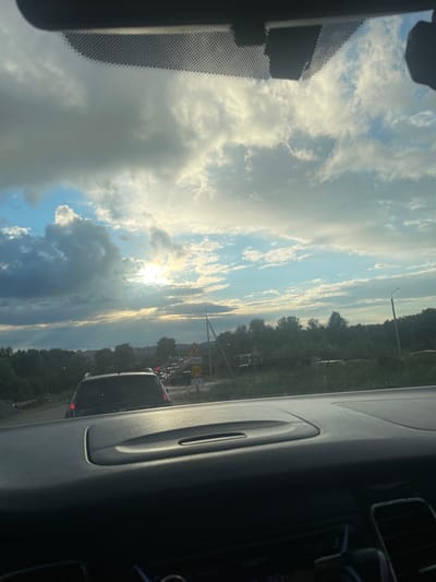 The image captures a scenic view from inside a vehicle, looking out through the windshield. The scene is dominated by a dramatic sky filled with a mixture of dark, heavy clouds and bright, sunlit patches, suggesting a dynamic weather pattern. The sun is partially obscured by the clouds, casting a warm, ethereal glow across the sky and illuminating the edges of the clouds with a golden hue.

In the distance, a treeline marks the edge of a landscape, likely rural or semi-rural, given the presence of vegetation. Several vehicles are visible on a road, indicating that the car the photo is taken from is in traffic or moving along a route. The presence of road signs suggests a developed area, though the overall impression is one of natural beauty contrasting with everyday infrastructure. One sign is partially visible, featuring a red circle with the number "30" inside, likely indicating a speed limit. Another sign says "СТОП," which means "STOP" in Russian.

The interior of the car is partially visible, with the dashboard and a portion of the passenger seat contributing to the frame. The perspective is from the driver's or front passenger's seat. The time of day appears to be late afternoon or early evening, given the angle of the sun and the quality of the light. The weather is clearly transitioning, with both cloudy and sunny elements present. The overall mood is one of quiet observation of the natural world from within the controlled environment of a vehicle, possibly during a commute or a journey. The location context of Gavrilovka, Russia, adds a specific geographical anchor to the scene.