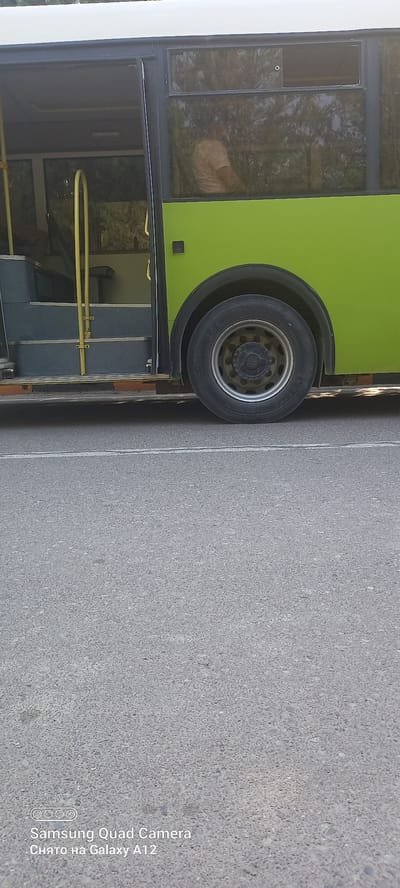 A partially visible, light-colored bus with a green lower body is parked on an asphalt road in Levakant, Tajikistan. The bus has its doors open, revealing a set of steps leading inside. A person wearing a light-colored t-shirt is visible through the tinted windows of the bus, seated and facing away from the camera. The front wheel of the bus, a large black tire with a silver rim, is prominent in the foreground. The road surface is asphalt with a white line painted on it. The environment appears to be outdoors, with trees and sky reflected in the bus windows. A watermark at the bottom of the image indicates it was taken with a Samsung Quad Camera on a Galaxy A12.