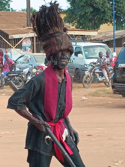 An older man with dark skin and a large headdress decorated with feathers stands on a dirt road. He wears a black shirt and pants with a wide red sash tied around his waist and over his shoulders. He holds a long, dark object that resembles a rifle. In the background, there are buildings, a white van, a motorcycle with two people on it, and a few other individuals. The setting appears to be an outdoor event or performance in a village or town.