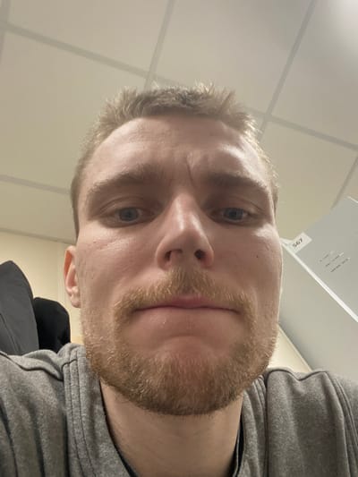 A direct, low-angle headshot captures a light-skinned adult male, presumably located in Nikol'skoe, Russia. The male has blue eyes and is looking directly at the camera. His short, light brown hair is disheveled on top. A blonde-to-light brown mustache and a short beard cover his upper lip, chin, and jawline. His forehead displays several horizontal lines. He is wearing a grey, possibly hooded, garment, visible around his neck and shoulders. The background consists of a light-colored dropped ceiling with a grid of square or rectangular acoustic panels. To the subject's left, a dark, indistinct fabric or object is partially visible. To his right, a light-colored vertical surface, possibly a cabinet or partition, displays the black numbers "567" and some smaller, illegible text. The scene is illuminated by overhead artificial lighting.