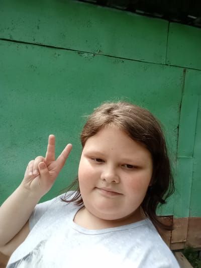 A young person is taking a selfie outdoors in front of a green, weathered wall. The person has brown hair and is wearing a light gray t-shirt. They are holding up two fingers in a peace sign. The wall appears to be made of metal panels that are peeling and discolored. The image is taken in the city of Pavlovsky Posad, Russia.