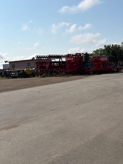 The image depicts an outdoor scene on a bright, sunny day in Bazan, Bulgaria, showcasing a collection of agricultural machinery. The main subject is a large, complex red farm implement, likely a seed drill or cultivator, occupying the left side of the frame on a paved surface. This prominent machine features multiple rows of discs or tines, a substantial seed hopper, and robust structural components. Visible text on its side clearly identifies the brand as "VADERSTAD" and likely the model "RAPID".

Behind this central piece of equipment, a variety of other farm machines, rendered in different colors including yellow and blue, are parked. Further back, a light brown or pinkish building with visible windows stands among green trees, suggesting a workshop, office, or storage facility. The foreground and right side of the image are dominated by a wide, light gray paved area, resembling an asphalt road or a large parking lot, showing slight texture and minor imperfections. Above, the sky is clear and blue with scattered white cumulus clouds. The scene conveys a sense of agricultural industry, possibly a dealership or a farm equipment storage yard. No people are visible, and there is no apparent activity other than the static presence of the machinery.