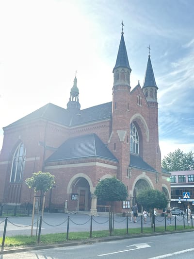 The image shows a red brick church with two spires topped with crosses. The church has arched windows and doorways, and a dark tiled roof. In front of the church, there is a paved area with a low chain fence. Two young trees on stakes are planted in front of the fence. Several people are visible in front of the church, some standing and some walking. To the right of the church, a modern building with windows is visible, along with a sign with a pedestrian symbol and text in Polish. A car is parked in front of the modern building. The sky is blue with some wispy clouds, and the sun is shining brightly, causing some lens flare in the image. The street in the foreground has a white arrow painted on it. The image is likely taken in Nowy Sącz, Poland, given the signage.