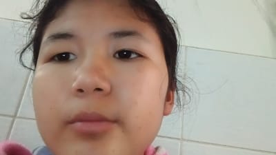 This is a selfie of a young East Asian female. She has dark hair, brown eyes, and fair skin with some freckles and blemishes. She is wearing a pink and white striped shirt. The background appears to be a tiled wall, possibly in a bathroom or kitchen. The image is not specific enough to determine a location within the city of Uchkun, Kyrgyzstan.