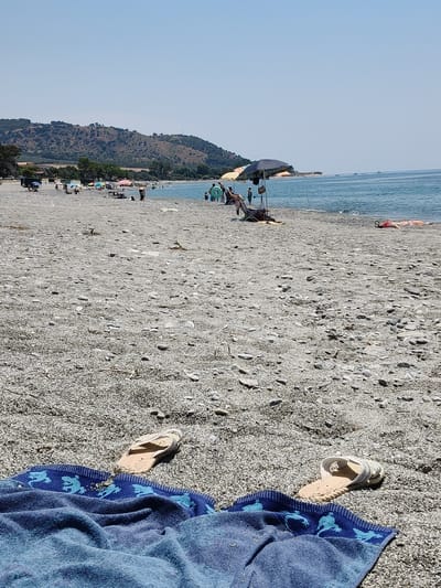 The image depicts a sunny day at a pebble beach in Bova Marina, Italy. In the foreground, a blue towel with a dragon pattern is spread out on the pebbles, with a single light-colored sandal placed on top of it. Another identical sandal is partially visible on the right side of the frame, also resting on the beach.
Further down the beach, several people are visible. Some are relaxing under umbrellas, while others are wading in the shallow, turquoise water. Children can be seen playing near the shoreline, and a few boats are anchored offshore, suggesting a recreational atmosphere. The beach itself is covered in smooth, grey pebbles, characteristic of the Mediterranean coastline.
Behind the beach, a gentle slope rises, covered in dry vegetation and scattered trees, leading up to a hazy, brown hillside under a clear, bright blue sky. The overall mood is relaxed and summery, indicative of a typical vacation scene. The time of day appears to be midday, given the strong sunlight and distinct shadows. There is no visible text in the image.