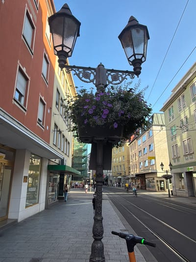 An eye-level, outdoor, slightly low-angle shot captures a street scene in Würzburg, Germany. The foreground is dominated by the ornate, dark metal post of a street lamp adorned with a hanging basket of purple flowers. Beside the lamp post, the handlebars of an orange and black electric scooter with a green illuminated display are visible.

The street runs alongside the lamp post and is paved with asphalt, featuring tram tracks. On the left side of the street, a sidewalk made of grey paving stones is lined with storefronts. The buildings on this side are predominantly two to four stories high. The closest building has a reddish-orange facade with white window frames and closed roll-down shutters. Further down, other buildings are painted in lighter colors, including yellow and a pale green. One building is partially covered in scaffolding and green netting, indicating construction or renovation.

On the right side of the street, buildings are also visible, painted in light yellow and pale green hues. These buildings also appear to be two to four stories tall and have multiple windows. The street is active with people: a woman in a black top and blue jeans walks away from the camera on the sidewalk. Further down, several people are seen on the sidewalk and a few individuals are cycling on the street. The sky above is a clear, bright blue, suggesting it is daytime and fair weather. The overall impression is a bustling urban streetscape in a European city.