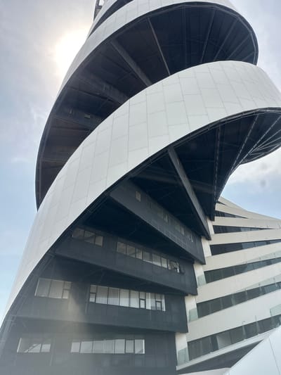 The image depicts a low-angle view of the Solara Medical Center in Tlaxcalancingo, Mexico. The building features a distinctive helical design, with white panels wrapping around a dark, angular core structure. Adjacent to the spiral section is a taller, more conventional building section with a pattern of white and black panels with glass windows. The sky is visible in the background with light clouds.