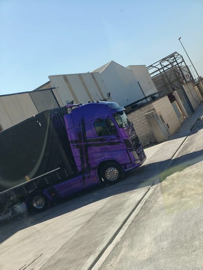 A vibrant purple semi-truck with a black canvas-covered trailer is the central subject of this image, captured in an industrial park within Polígon Industrial Dels Vents, Spain. The truck's cab features a distinctive black abstract graphic design and polished chrome wheels.
The setting consists of various industrial buildings, primarily featuring corrugated metal siding in shades of grey and white, under a clear, bright blue sky. Some structures appear to be warehouses or factory units. In the background, there is also the skeletal metal framework of what might be a partially constructed or deconstructed building or sign, and a tall street light pole. Lower walls constructed from light-colored brick are also visible, along with concrete utility boxes and other industrial elements.
The truck is situated on a paved asphalt and concrete lot or road, with strong, defined shadows cast by objects, indicating bright sunlight and a clear day, likely during mid-morning or afternoon. There isn't clear activity, but the truck appears to be parked or moving slowly within the industrial environment.
A notable detail is a small, rectangular yellow sticker affixed to the side of the black trailer, prominently displaying the word "ATTENTION" above a black symbol and some unreadable smaller text. The image is captured from an angled perspective, giving a dynamic feel to the industrial scene.