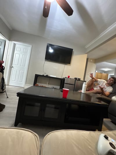 This image depicts an interior residential scene, likely a living room, in a structure located in the city of South Bend, United States; however, no specific geographic identifiers within South Bend are visible in the photograph.
The environment features light-colored walls, a white ceiling with a dark brown three-bladed ceiling fan and light fixture, and light-toned flooring. A white five-panel interior door is visible on the left. In the background right, an archway opens to another room, possibly a kitchen or dining area, with light-colored wooden cabinets and a ceiling light visible.
A single adult male is positioned on the right side of the image, reclining on a dark-colored sofa. He has short, dark hair and is smiling, looking towards the camera. He is wearing a white t-shirt and red shorts. His legs are bent, with his left foot resting on his right knee. His left hand is raised, holding a small, light-colored object between his thumb and index finger, with his remaining fingers splayed.
In the foreground, two light-colored upholstered sofa sections are partially visible, suggesting the vantage point of the photographer. A white Xbox controller rests on the right-most of these sofa sections.
A large, rectangular black coffee table occupies the center midground, featuring two red plastic cups and a white water bottle or tumbler on its surface. The table has two glass-fronted cabinets below. Behind the coffee table, against the wall, is a narrow black console table holding a white electronic device, possibly a gaming console. Above this console, a large, black flat-screen television is mounted on the wall; it is powered off, reflecting ambient light. To the right of the console table, a two-section compact refrigeration unit is visible. To the far left, partially obscured, is a golf bag containing golf clubs, supported by a stand, next to a red object.