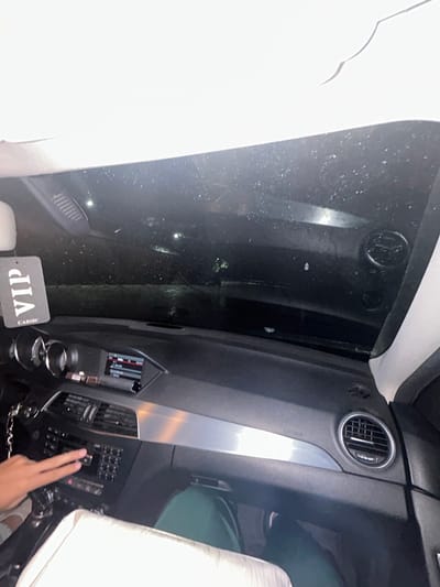 The image captures the interior of a vehicle, likely a luxury car or a transport van, at night in Dolna banya, Bulgaria. The perspective is from the passenger side, looking towards the driver's area and out the front windshield.
The main subjects include the vehicle's dashboard, a person's hand, and the exterior visible through the windshield. The dashboard is dark grey or black, featuring multiple circular air vents with metallic rings, a prominent silver trim piece running vertically, and an intricate control panel. A hand, likely belonging to the driver or a front-seat passenger, is shown pressing buttons on this control panel.
On the left side of the frame, a large portion of light-colored upholstery or seat material is visible, contrasting with the dark interior surfaces. Another light-colored seat or headrest is partially visible on the far right.
Through the windshield, the outside environment is very dark, indicating nighttime. Numerous small, bright specks are scattered across the windshield, suggesting water droplets from rain or heavy condensation. Faint, blurry lights are discernible outside, possibly from streetlights or distant vehicles.
A notable detail is a black rectangular tag hanging in the lower-left portion of the dashboard area, displaying the white, inverted text "VIP CABIN". The overall scene suggests a journey or a parked vehicle during dark hours. No specific emotions are evident.