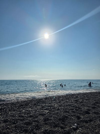 This image depicts a sun-drenched day at a pebble beach with a group of people swimming in the clear blue sea. The sun is high in the sky, casting a bright glare on the water's surface and creating a visible sunbeam effect in the sky. The foreground is dominated by dark grey and black pebbles, with a few lighter-colored stones scattered amongst them. Gentle waves are breaking on the shore. In the water, several individuals are visible, some with their heads above water, enjoying the sea. The horizon is sharp, separating the calm sea from the clear blue sky. The overall scene conveys a sense of leisure and a warm summer day. The location is identified as a beach in the city of Progress, Russia.