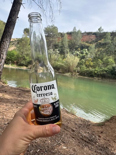 A right-handed person holds a Corona beer bottle against the backdrop of the Salto de la Novia waterfall in Jérica, Spain. The bottle is almost empty, with only a small amount of beer remaining. The bottle is angled slightly upwards, with the label visible. The backdrop includes a green body of water, likely the river, and the rocky terrain of the waterfall area covered with trees and bushes. The sky is overcast. A tree branch is visible in the lower left.