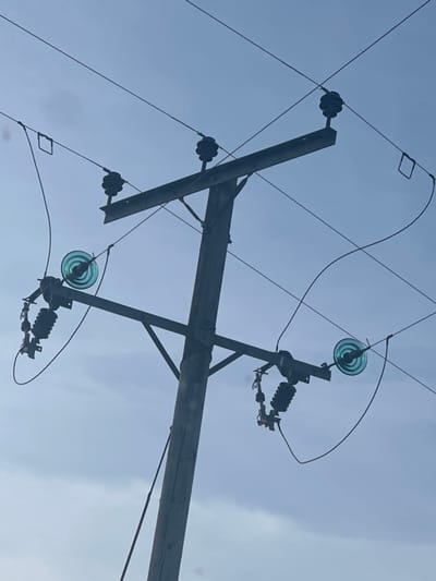 The image depicts a utility pole situated in Fowlis Easter, United Kingdom, viewed from a low angle against a clear, light blue sky with subtle cloud formations. The pole is constructed of wood or concrete, stained dark, and extends vertically upwards. Two horizontal crossarms are affixed to the pole; an upper, shorter crossarm supports two dark-colored, multi-skirted pin-type insulators, each carrying an electrical line. A lower, longer crossarm, braced by a diagonal support, features a light blue/green glass disc insulator on each end, which connect to open-style fuse cutouts. Multiple dark electrical lines traverse the frame, supported by the various insulators, indicating an active electrical distribution system. No individuals are present in the scene. The overall environment suggests an outdoor utility installation during daylight hours.