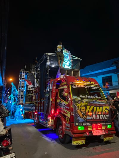 A customized red medium-duty truck, extensively modified into a mobile sound system, is depicted at night on a paved street in Kedungbanteng, Indonesia. The truck's cab and body are adorned with "KING AUDIO" branding, including a prominent crowned lion logo, multiple decals such as "Ferrari" and "Dwi Putra Audio," and numerous red, blue, amber, and green LED lights. The Indonesian license plate "P 8560 GG" is visible on the front bumper.

Mounted on the truck bed is a multi-tiered sound system structure composed of speakers, amplifiers, and lighting rigs. A male subject, wearing a patterned long-sleeve shirt and shorts, stands atop the highest accessible tier of this structure, facing away from the viewer, next to a large, illuminated lion head graphic. Another male subject is partially visible further back on a lower tier, operating equipment. An Indonesian flag is displayed on the side of the sound system.

In the background, to the left of the truck, a group of people is visible, illuminated by blue stage lighting. On the far left, a streetlight is present, and a network of overhead wires descends vertically. A partially visible motorbike is in the immediate foreground on the left. To the right of the truck, a building with a blue upper floor is visible. The overall scene suggests a public event or parade under artificial illumination.
