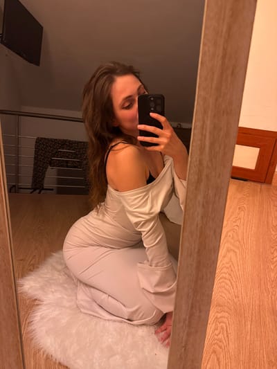 An image taken in Cava de' Tirreni, Italy, depicts a female individual captured in a mirror selfie within an indoor setting. The individual, possessing long brown hair, is shown in a kneeling or crouched position on a white, fluffy rug. She is wearing a light-colored, possibly beige or off-white, long-sleeved garment with an off-the-shoulder design, revealing a black bra strap on her left side. Her left hand holds a black smartphone, equipped with a pop socket, positioned to take the photograph.
The mirror, framed in light wood, reflects the interior of what appears to be a private residence. Visible in the background reflection are a black flat-screen television mounted high on a wall, a metal railing (suggesting a loft or upper level), and a dark patterned garment draped over the railing. To the right, a brown wooden picture frame leans against a wall on a light wood-grain floor. The overall lighting is warm-toned.