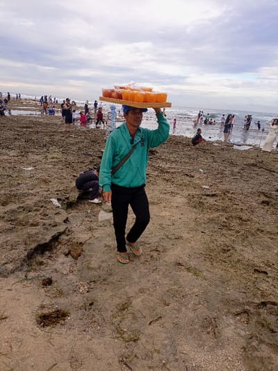 A man, wearing a turquoise shirt, black pants, and sandals, is walking on a sandy beach with a tray of orange drinks balanced on his head. He is facing the right side of the image, with a group of people in the background. There are other people, some sitting and some standing, in the background. The man appears to be walking towards the ocean, which is visible in the background. The sandy beach is mostly bare, with some patches of vegetation. The coordinates provided locate the scene at a beach in Pameungpeuk, Indonesia.