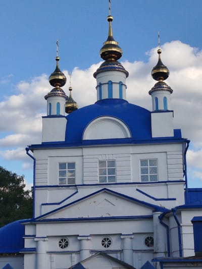 The image shows the Dormition Church in Mamontovo, Russia. The church features white walls and blue roofs and trim. It has four golden onion domes with crosses on top. There are three visible windows on the main structure and smaller circular windows below. A tree is visible on the left, and the sky is partly cloudy.