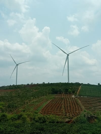 Two wind turbines are situated on a rolling hillside in Gao, Vietnam. The landscape consists of cultivated land, featuring rows of crops and varying vegetation. The sky is lightly clouded.