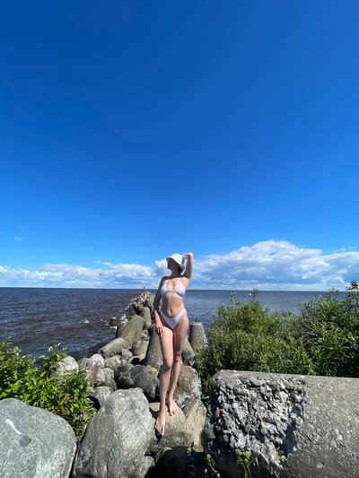 A woman in a white bikini and a white bucket hat stands on a rocky breakwater under a bright blue sky with scattered clouds. The breakwater extends into dark, choppy water. Green foliage grows on the sides of the breakwater. The woman is facing away from the camera, with her right arm raised, touching her hat. The scene appears to be a coastal area.