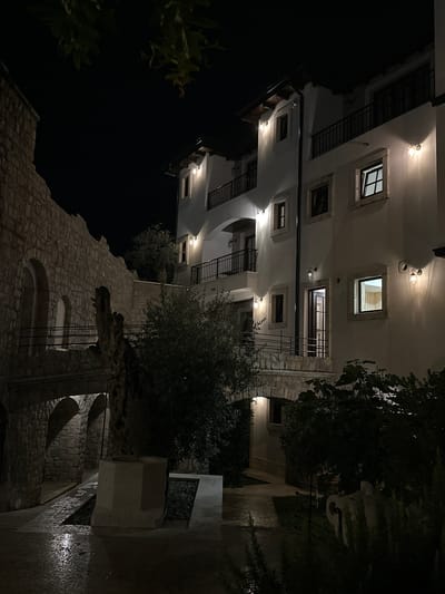In the city of Bartula, Montenegro, at night, a multi-story white building with arched windows and balconies is illuminated by exterior lights. Adjacent to it is a stone wall with arched openings. A stone bridge connects the wall to the building. In the foreground, a polygonal stone structure sits next to a dark green bush. The ground is wet, reflecting the light, suggesting recent rain. The sky is dark and clear, with leaves from an overhead tree visible in the upper portion of the image.