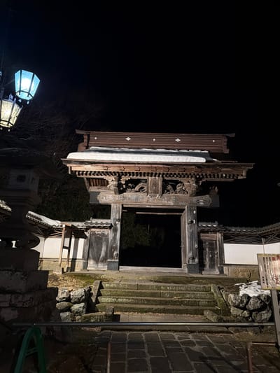 The image displays a traditional Japanese wooden gate at night in the Kanbayashi Onsen area of Yamanouchi, Nagano Prefecture, Japan. The scene is illuminated by artificial light sources, casting strong shadows against the dark background.

At the center, a multi-tiered wooden gate with intricate carvings and a dark brown tiled roof is visible. A layer of snow rests on the roof's upper tiers. The large central gate door on the right is partially open, revealing complete darkness within. Stone steps, partially covered in moss, lead up to the gate entrance from a paved stone pathway in the foreground.

To the left, a tall, multi-tiered stone lantern emits a bright, cool blue-white light, with a second, shorter lantern behind it showing a warmer, yellow-white glow. Patches of snow are present on the ground and surrounding low walls. The gate is flanked by traditional white walls with tiled roofs, featuring smaller wooden access doors on either side. Bare tree branches are discernible in the dark background. A warning sign or information panel is partially visible on the far right, near a small accumulation of snow on rocks. No individuals are present in the scene.