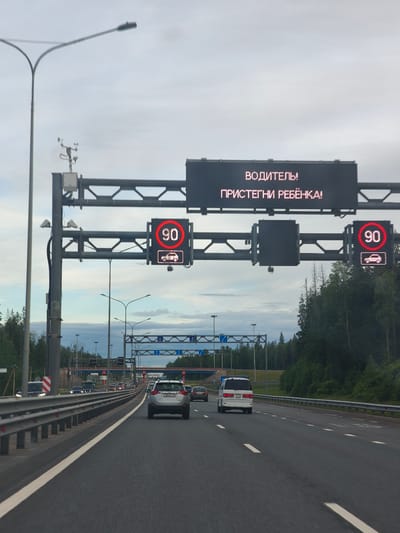 The image depicts a highway scene in Kissolovo, Russia, under an overcast sky, suggesting either early morning or a cloudy day. The focus is on a digital road sign displaying a message in Russian: "ВОДИТЕЛЬ! ПРИСТЕГНИ РЕБЁНКА!" which translates to "DRIVER! BUCKLE UP YOUR CHILD!". Below the text, there are two speed limit signs, each indicating "90" within a red circle, accompanied by an icon of a car.

The road is a multi-lane highway, with several vehicles visible. In the foreground, a silver SUV is in the right lane, while a white minivan is in the left lane, also traveling forward. Other cars are present further down the road. The setting is a highway bordered by a metal guardrail on the left and a dense line of trees on the right. Overhead, a large gantry supports the digital sign and other traffic management equipment, including what appear to be cameras and meteorological sensors. The overall mood is cautionary, with the digital sign serving as a reminder for child safety.
