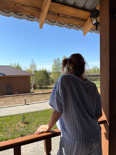 A woman stands on a wooden structure, possibly a porch or balcony, with a view of a landscape. The scene appears to be located in Kamennoye, Russia, based on the metadata. The woman, wearing a striped blue and white shirt, leans against the railing. She has her hair pulled back. The view includes a building with a dark roof, a walkway, grass, and trees under a bright blue sky. The wooden structure has visible beams and a light fixture.