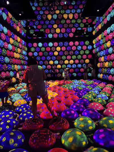 The image depicts an immersive art installation at teamLab Planets in Koto, Tokyo, Japan. The space is filled with numerous rounded, dome-like structures of varying sizes, densely packed together, creating a floor that resembles a landscape of oversized, colorful mushrooms or stones. These structures are illuminated with vibrant, glowing patterns of light in shades of blue, purple, green, yellow, and red, with some exhibiting polka-dot designs. The walls and ceiling of the room are also covered with a grid of similar glowing circular or ovular elements, creating a highly saturated and visually stimulating environment.

In the foreground and middle ground, several individuals are present. A person in the middle foreground, facing away from the camera, wears a light-colored shirt and shorts and is standing on one of the glowing structures. To the left, a child in a blue skirt and pink top is also interacting with the illuminated floor. Further back, other figures, including children and adults, are visible interacting with the art installation, with some appearing to walk or stand on the structures. The lighting is dramatic and theatrical, with the illuminated elements casting colored reflections onto the people and the surfaces. The overall impression is one of a surreal, futuristic, and interactive art experience.