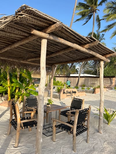 This image depicts a rustic and inviting tropical outdoor lounge area, likely a beach bar or restaurant, set in Kiwengwa, Tanzania.
The main subject is a large, open-sided structure with a thick, textured thatched roof, supported by sturdy, natural wooden posts. Underneath this shelter, several tables and chairs are arranged on the sandy ground. The chairs feature natural wooden frames with black woven or strapped seats, complementing the informal, beach-style decor.
Surrounding the seating area are vibrant green tropical plants, including prominent palm trees reaching towards a clear, bright blue sky. The ground throughout the visible area is light-colored sand.
In the mid-ground, a distinctive sign made of a light-colored, possibly carved stone or wood, reads "The Beach Lounge" and "Zanzibar" in a stylish script. Further in the background, partially obscured by foliage and structural elements, a person wearing a green shirt can be seen. Another building or structure made of natural materials is visible behind them, with text that appears to include "DURDSTONE HOTEL" (partially visible).
The bright natural lighting and clear sky indicate it is daytime, likely morning or late afternoon, with sunny and pleasant weather. The scene conveys a relaxed, tranquil, and tropical atmosphere.