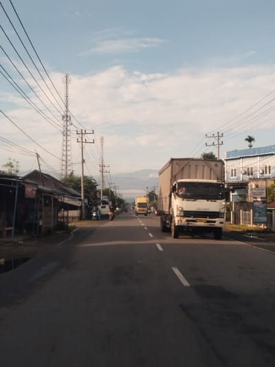 The image depicts a street scene in Lada, Indonesia, likely during daytime given the clear sky and shadows. The perspective is from a moving vehicle, looking forward along a two-lane asphalt road. On the left side of the road, a row of small, weathered shops with open fronts and simple roofing are visible. Utility poles with numerous wires are prominent throughout the scene, alongside tall cell towers in the distance.

Several vehicles are present. A white Isuzu truck with a large cargo container is the most prominent subject, driving in the right lane, heading away from the viewer. Another yellow truck is visible further down the road in the left lane, also moving away. Several motorcycles are also on the road, some with riders, indicating local transportation.

In the background, a blue and white building with air conditioning units is situated on the right side of the road, with signage indicating "CV. TAYTIHA JUAL TIKET PESAWAT" (CV. Taytiha Sells Plane Tickets) and "BSI". The landscape behind the buildings includes hills or mountains under a sky with scattered white clouds. The overall impression is a commercial or semi-urban area with active road traffic.