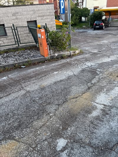 This media file captures an outdoor scene, likely a parking area or service road, in Camerino, Italy. The dominant feature is a heavily worn asphalt pavement covering most of the frame, characterized by numerous cracks and visible white and grey aggregate. Patches of yellowish staining are also present on the road surface.

To the left, a light-colored brick building stands, next to which an orange barrier control box is visible. This box features the "FAAC" logo and some indiscernible text or symbols. A green metal gate structure is associated with this barrier system, suggesting a controlled access point. Along the curb next to the asphalt, sparse weeds and a small piece of crumpled litter are visible.

Further into the background on the left, a black car is parked near a blue sign displaying a prominent white "P", indicating a parking zone. Beyond the car and the sign, more buildings with yellow awnings and dense green foliage from trees and bushes can be seen.

The scene appears static, with no people visible and no apparent activity taking place. The lighting suggests daytime, possibly under overcast conditions, as shadows are soft and diffused.