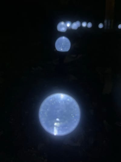 The image captures a nighttime scene in Branipole, Bulgaria. The primary subjects are a series of glowing, orb-like lights. These lights appear to be arranged in a loosely diagonal line, moving from the upper left to the lower right of the frame. The lights themselves are a bright, bluish-white and are slightly blurred, suggesting they might be small LED lights or similar illuminations.

In the foreground, on the right side of the image, a larger, more distinct light orb is visible. It is the brightest and most detailed of the lights, showing some internal texture or reflections. To the left of this large orb, a smaller, similar orb is also present, appearing slightly further away. Further to the left and slightly higher in the frame, a chain of even smaller, more numerous orbs is visible, receding into the darkness.

The background is almost entirely black, indicating a dark, likely outdoor setting at night. There are some faint, indistinct shapes and textures visible in the background, suggesting a natural environment such as foliage or uneven ground, but they are too blurred to be clearly identified. The overall mood is somewhat mysterious and serene due to the dark setting and the gentle glow of the lights. No people are visible in the image. The time of day is clearly nighttime. There are no visible text elements in the image.