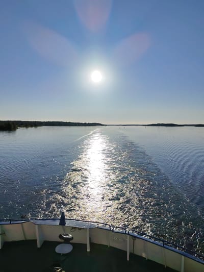 The image presents a detailed view from the stern of a moving vessel on a wide, calm inland waterway, likely near Goritsy, Russia, on the Sheksna River or Volga-Baltic Waterway. The scene is illuminated by a bright, undiffused sun, visible as a distinct circular disc in the upper-right quadrant of a clear, light blue sky, creating a pronounced starburst lens flare and additional diffuse light artifacts.
The sun's direct reflection forms a brilliant, shimmering path on the water, precisely aligned with the turbulent wake trailing directly behind the vessel. This wake, characterized by agitated white and sparkling water, extends from the immediate foreground into the mid-distance. On either side of the wake, the water surface exhibits gentle, uniform ripples, reflecting the clear sky.
Distant, low-lying forested landmasses or shorelines are visible on both the left and right sides, forming a dark, relatively flat horizon line across the mid-ground. Several small, dark navigational buoys are discernable on the water's surface in the mid-distance, indicating a navigable channel.
In the immediate foreground, the stern of the viewing vessel is partially visible, featuring a curved white superstructure topped with a blue railing. On the deck to the left, a small, round white table-like object with a dark cylindrical base, and a larger dark cylindrical object (possibly a covered mooring bitt), are present. A similar dark cylindrical object is visible on the right side of the deck. No individuals are depicted in the scene.