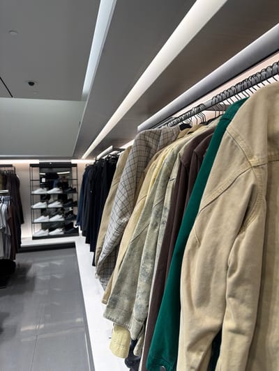 A close-up perspective reveals a clothing rack overflowing with jackets and shirts. The garments, stacked tightly together, feature a range of colors including beige, green, brown, and a checkered pattern. Below, dark-colored shirts and pants are also visible. In the background, a display showcases several white shoes. The overall impression is a well-stocked retail environment.