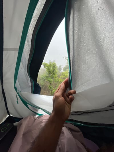 The image is a point-of-view shot from inside a tent, looking out through an partially open zipper. Raindrops are visible on the tent fabric, suggesting a wet environment. Outside the tent, a lush green forest with trees and foliage can be seen under an overcast sky. A hand is reaching out to adjust the zipper. The tent interior shows a pink blanket and a black object with a green light. The scene appears to be in a natural, outdoor setting, likely a campsite, given the presence of the tent and the surrounding forest. The location is identified as Unknown, India.