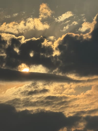 The image depicts a sky filled with clouds during what appears to be sunset or sunrise. The sun is partially obscured by dark clouds, creating a halo effect and casting a warm, golden light on some of the clouds. The lighting suggests dramatic atmospheric conditions. The image is a close-up of the sky and does not contain any elements that would indicate a specific location within the city of Constanța, Romania, or any other geographical context. There are no people, subjects, objects, or actions visible in the scene.