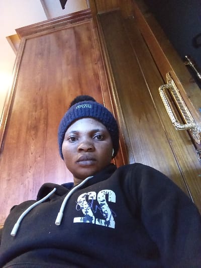 An adult female is depicted in a close-up, slightly low-angle indoor shot. She wears a dark blue knit beanie with a black pom-pom and a white embroidered logo on the front, along with a black hooded sweatshirt featuring white drawstrings and a white graphic print on the chest. A white wireless earbud is visible in her right ear, and she has a small silver nose piercing on her right nostril. Her gaze is direct and her expression neutral. The background consists of polished reddish-brown wooden paneling or a door. To the right, an ornate gold-colored metal handle is partially visible. A portion of a light-colored ceiling with a dark beam is present at the top. The specific location within the city of Madinaty, Egypt, is not discernible from this indoor scene.
