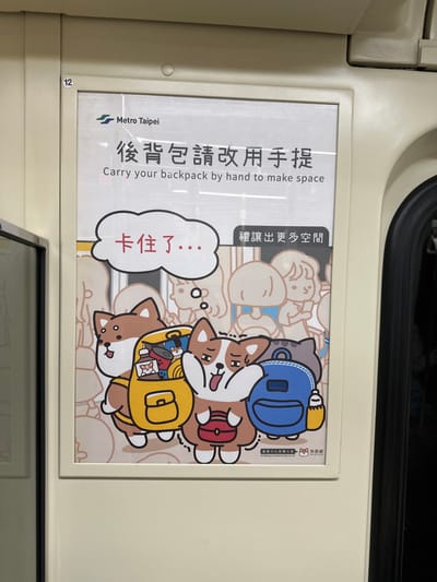 A rectangular public service announcement poster, framed in light beige, is affixed to a cream-colored interior wall panel of a Metro Taipei train car, located within New Taipei, Taiwan. The poster features the "Metro Taipei" logo in the upper left. Its primary message, in large Chinese characters "後背包請改用手提," is translated below as "Carry your backpack by hand to make space."
The lower portion of the poster displays a colorful cartoon illustration against a background of faint, stylized outlines of human figures, representing a crowded environment. In the foreground, three anthropomorphic animals are depicted. On the left, a Shiba Inu wears an open, overflowing yellow backpack containing various items, including bottles and small cat figurines. In the center, a Corgi exhibits a distressed expression with an open mouth and raised paws, holding a small red handbag. A white speech bubble above the Corgi contains red Chinese characters "卡住了...", signifying "Stuck!" or "Jammed!". To the right, a grey cat is partially obscured by the Corgi, wearing a blue backpack with an attached yellow bottle. A smaller box on the right side of the poster contains Chinese text "禮讓出更多空間," which translates to "Be courteous and make more space."
The surrounding environment consists of the train car's light beige interior, with a faint number "12" visible above the poster frame on the upper left. A dark vertical element, likely a window or door frame, is present on the right edge of the image. The scene is evenly illuminated by artificial overhead lighting.