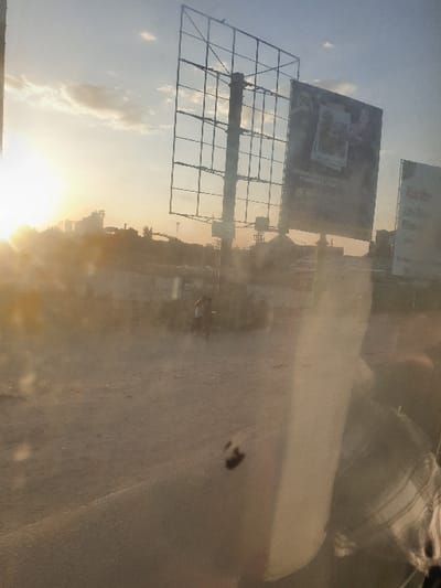A low-angle shot captures a partly cloudy sky at sunset. Silhouetted billboards stand tall, their metal frames visible. A hazy atmosphere obscures details of the background. The scene seems to be viewed through a grimy window, distorting the light and creating a smeared effect. The sun casts a warm glow, emphasizing the billboards' forms.